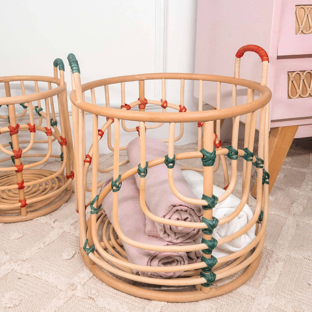 Close-up of the larger handcrafted Iris baskets set by MOMIJI in natural rattan with red and green bindings filled with rolled towels, with the smaller basket beside a cabinet.