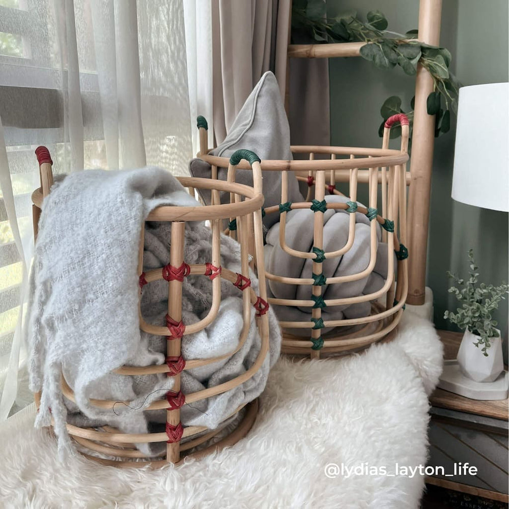 Handcrafted Iris baskets set by MOMIJI in natural rattan with red and green bindings arranged on a bed, filled with blankets and cushions in a softly lit bedroom.