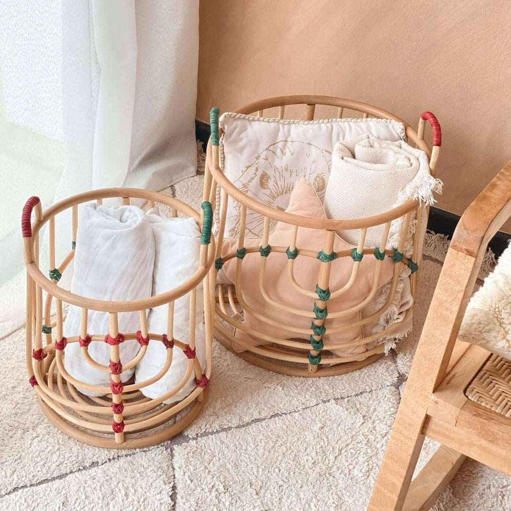 Handcrafted Iris baskets set by MOMIJI in natural rattan with red and green bindings placed side by side, filled with folded blankets on a rug in a room with a wooden chair.