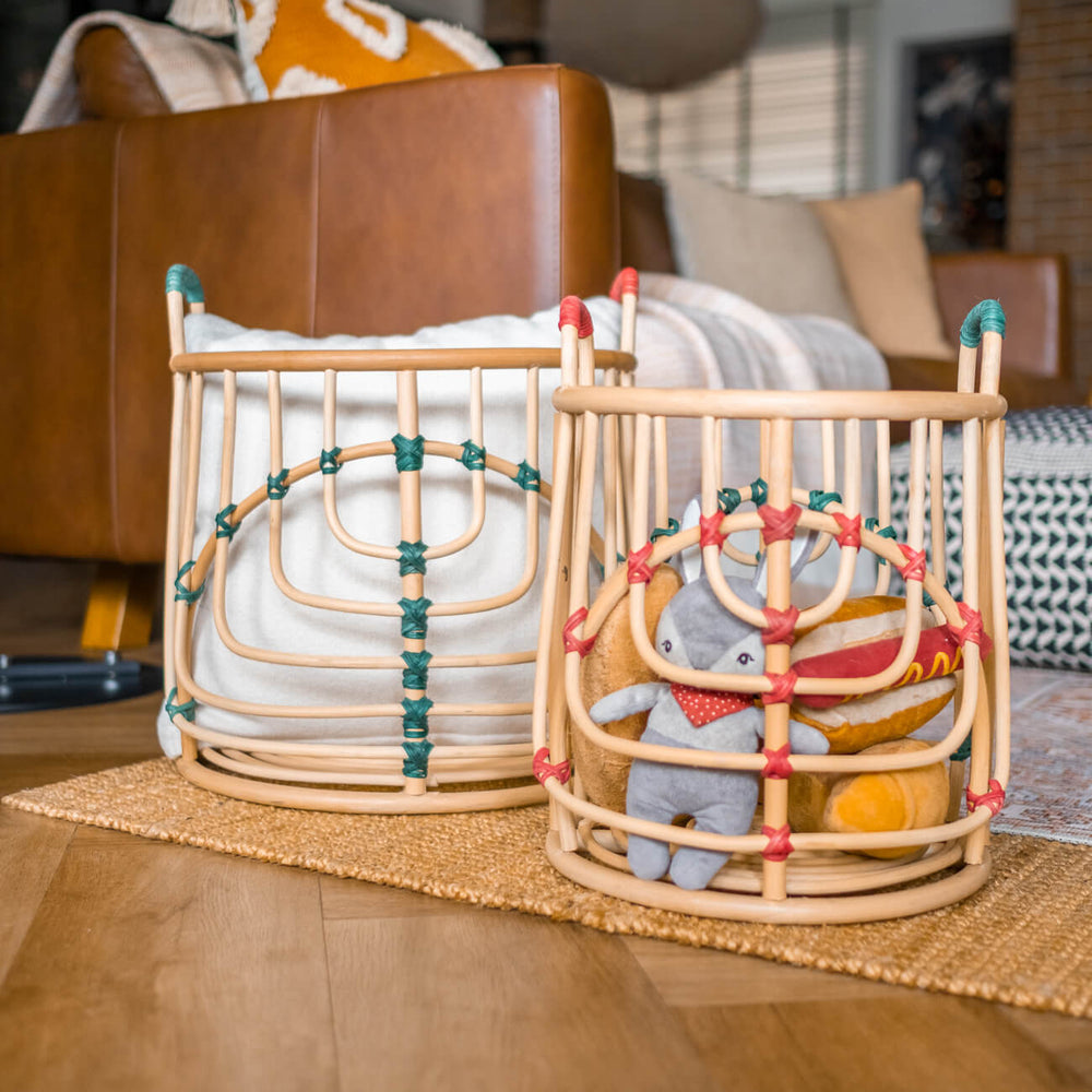 Handcrafted rattan wicker Two Iris baskets by MOMIJI placed side by side, filled with cushions and plush toys placed side by side near a sofa, in natural rattan with green and red bindings.