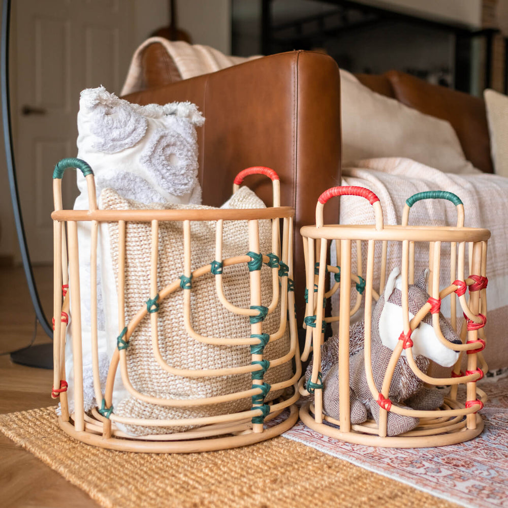 Side view of two handcrafted rattan Iris baskets by MOMIJI filled with cushions and toys in natural rattan with green and red bindings, placed side by side near a sofa.