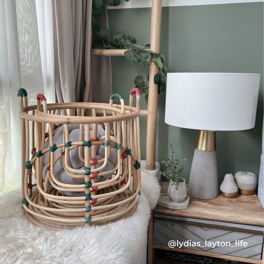 Stacked handcrafted Iris baskets set by MOMIJI in natural rattan with red and green binding, filled with yarn balls in a cozy room with a bedside lamp.