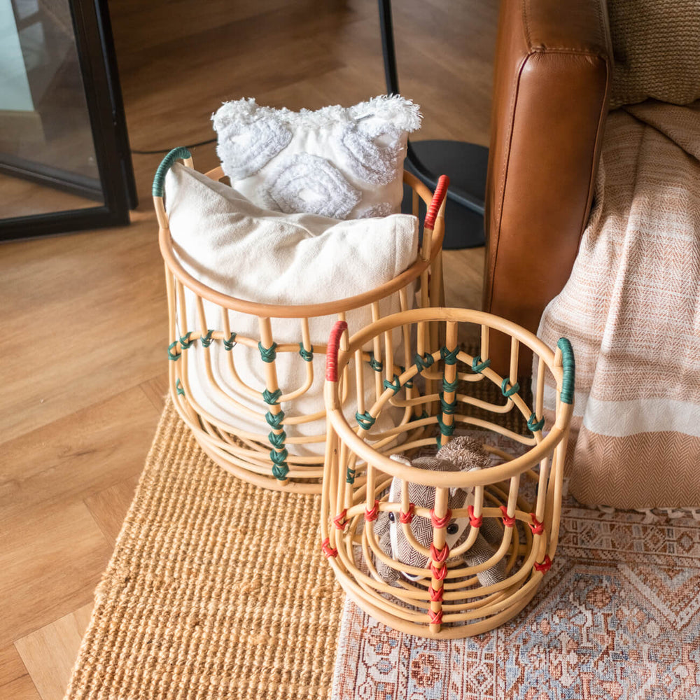 Handcrafted Iris baskets set by MOMIJI in natural rattan with red and green bindings, filled with plush toys and cushions on a rug with a sofa behind.
