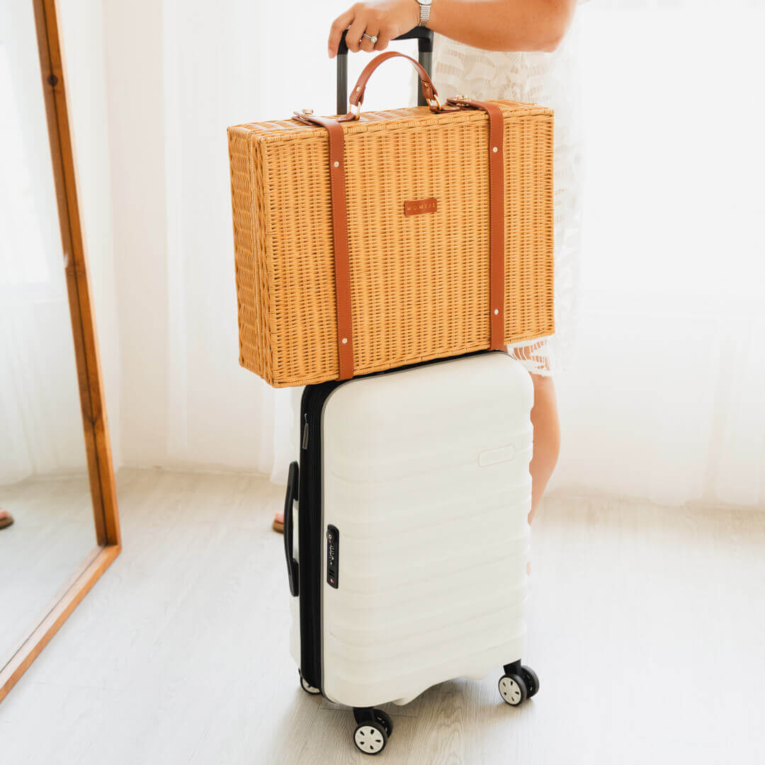 Woman holding a natural Jasmine rattan Suitcase by MOMIJI on top of a white suitcase in a minimal room setting.