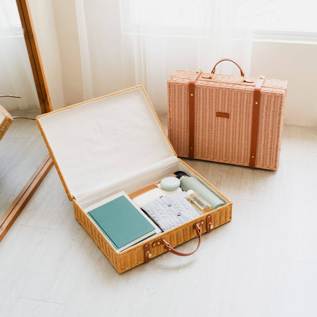 Opened natural version of Jasmine rattan Suitcase by MOMIJI showing clothes, books, and daily essentials inside, placed on the floor with an extra pink version beside it.