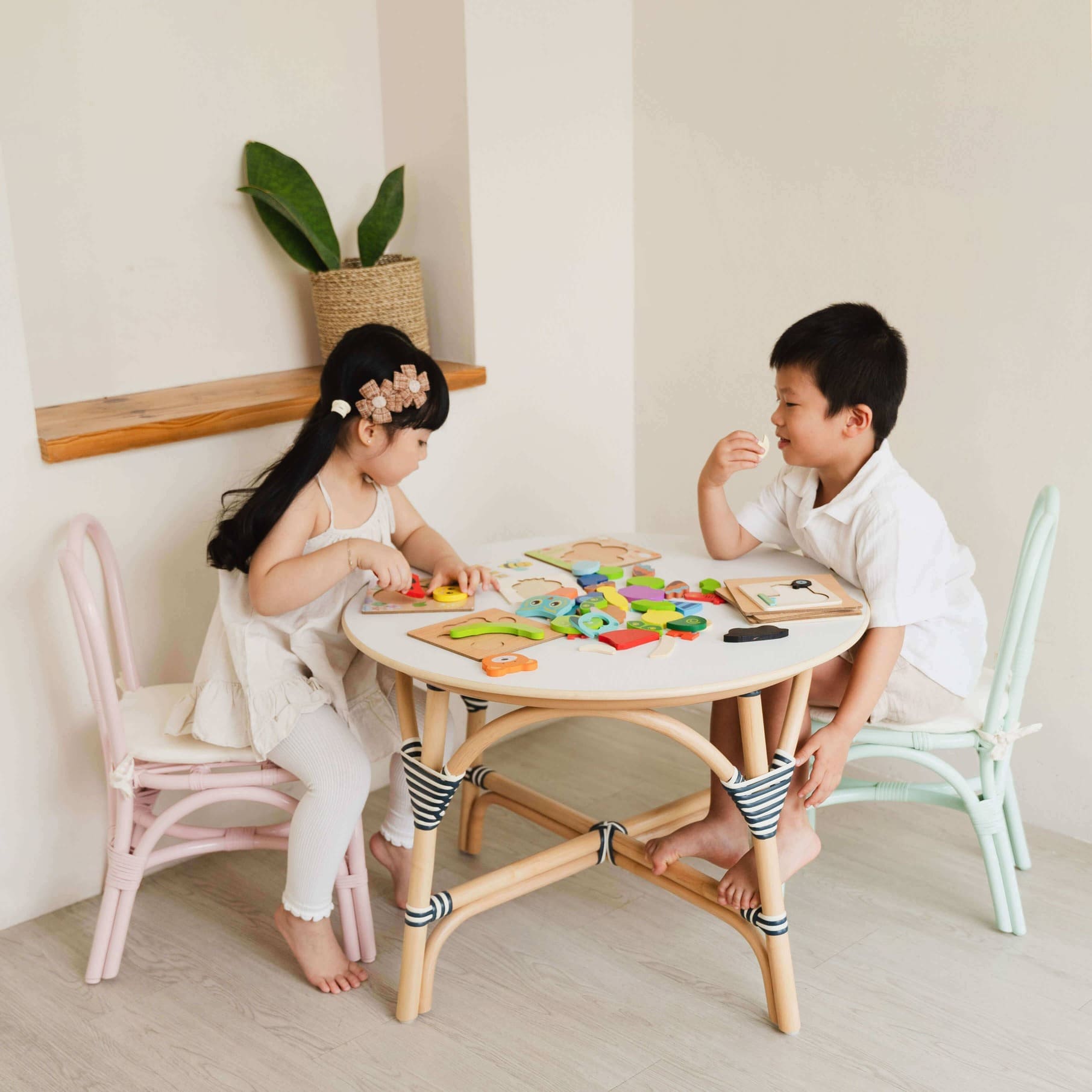 Two children sit in Bunny Rattan Chairs Pink & Mint-Green by MOMIJI playing with toys at a small round table in a bright room.