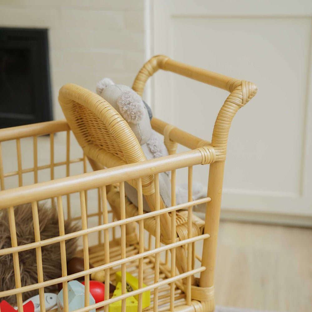 Side view of a handcrafted wicker rattan Kids Shopping Cart by MOMIJI filled with toys and a doll in the baby seat