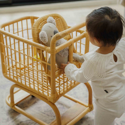 Child placing a doll in the baby seat of a handcrafted wicker rattan Kids Shopping Cart by MOMIJI, highlighting the 2 in 1 stroller feature