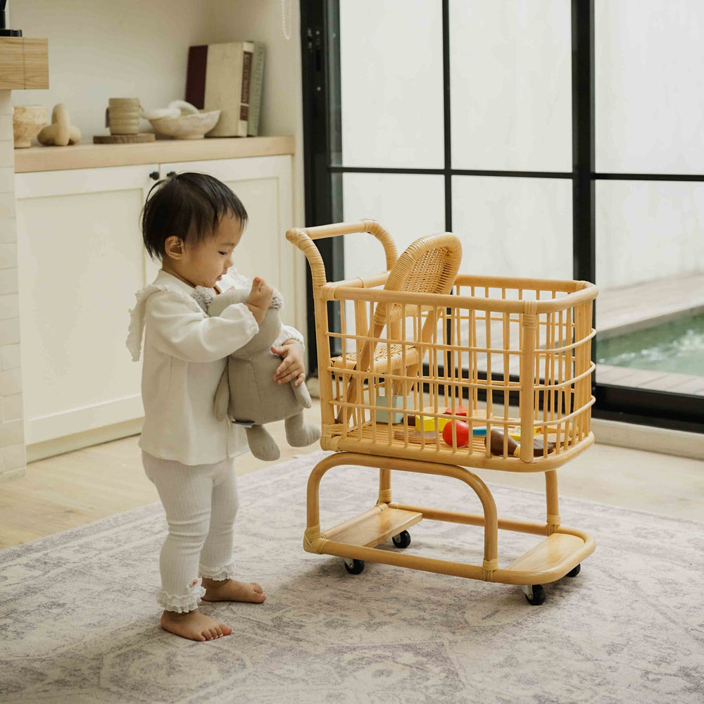 Child playing with a handcrafted wicker rattan Kids Shopping Cart by MOMIJI filled with toys in a bright room with large windows 