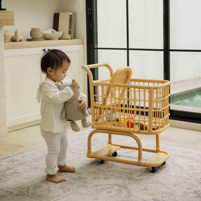 Child playing with a handcrafted wicker rattan Kids Shopping Cart by MOMIJI filled with toys in a bright room with large windows 