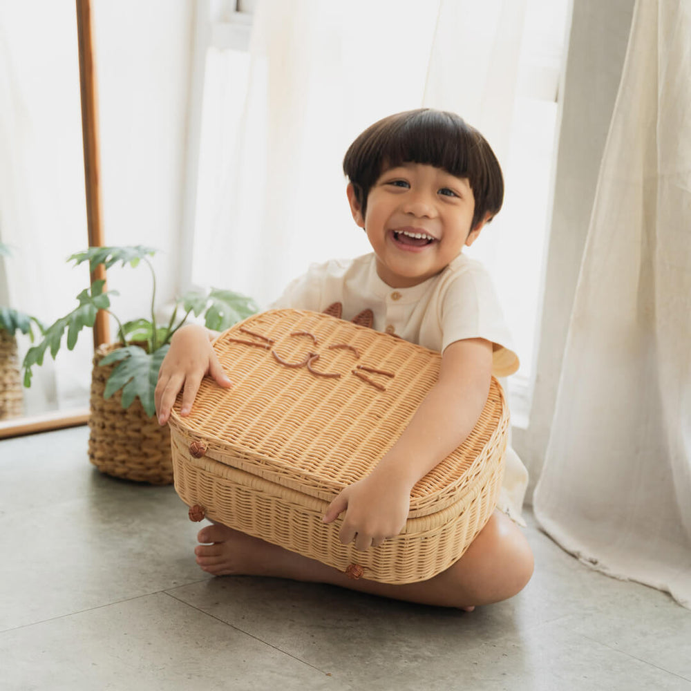 Smiling child sitting on the floor while holding the handcrafted rattan wicker Kitty Kids Suitcase by MOMIJI.