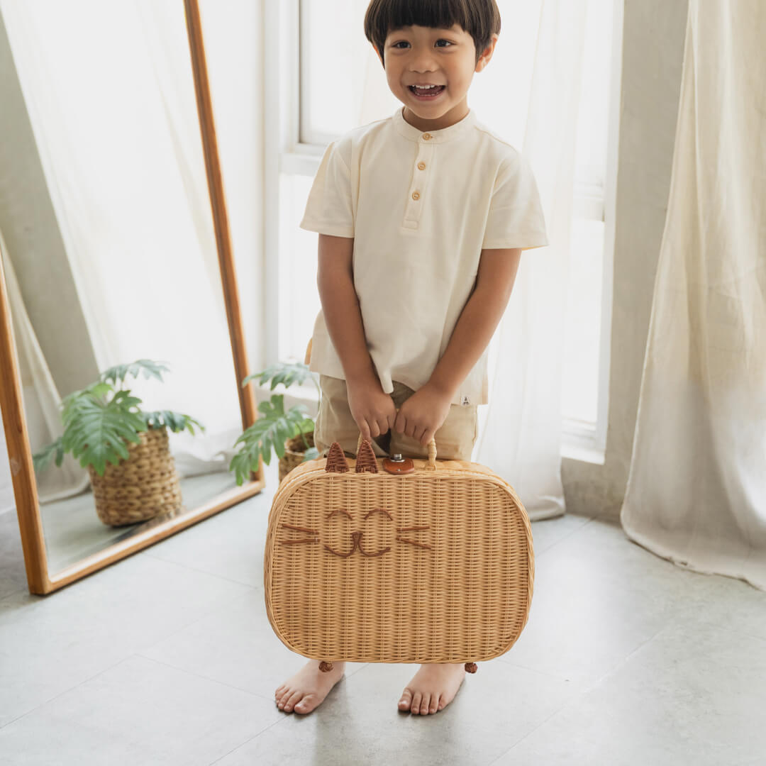 Happy child holding the handcrafted rattan wicker Kitty Kids Suitcase by MOMIJI in a room with plants and a mirror.