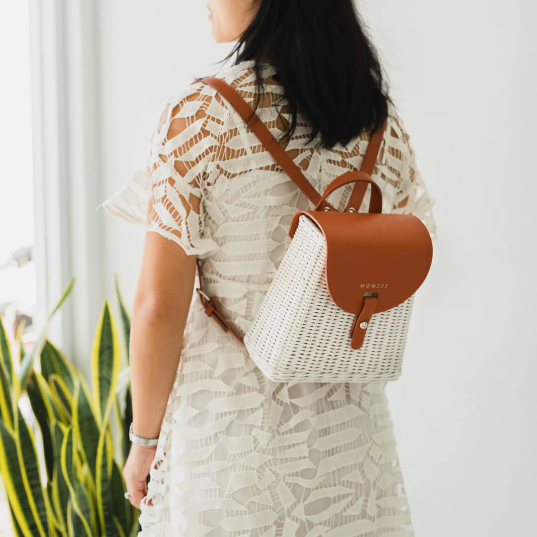 Woman wearing the handcrafted rattan Layla Backpack by MOMIJI in white, featuring a brown leather flap and adjustable straps.