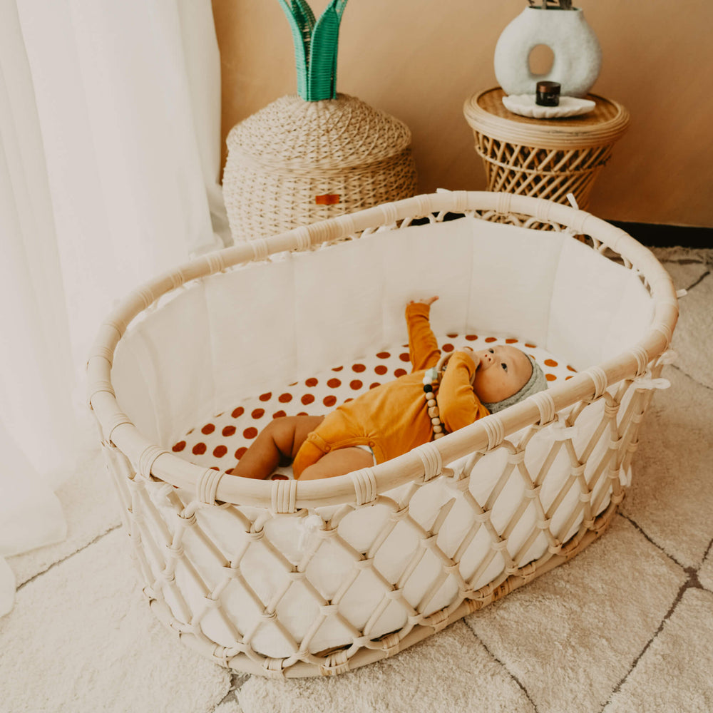 Baby resting in the Layla Bassinet by MOMIJI with a polka dot sheet in a cozy room with rattan basket and stool.
