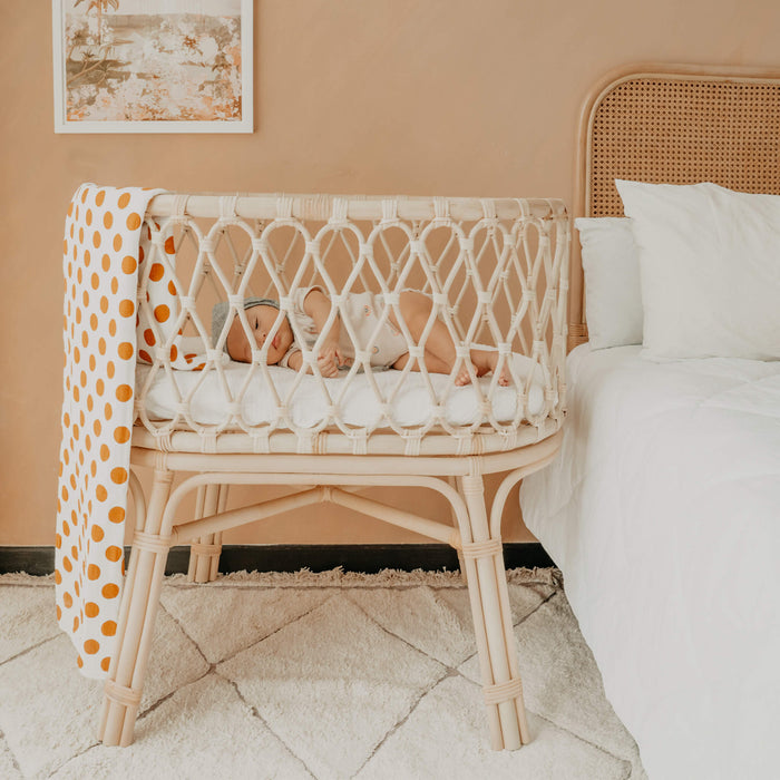 Baby sleeping in the handcrafted rattan wicker Layla Bassinet by MOMIJI with a polka dot blanket beside a bed in a bedroom.