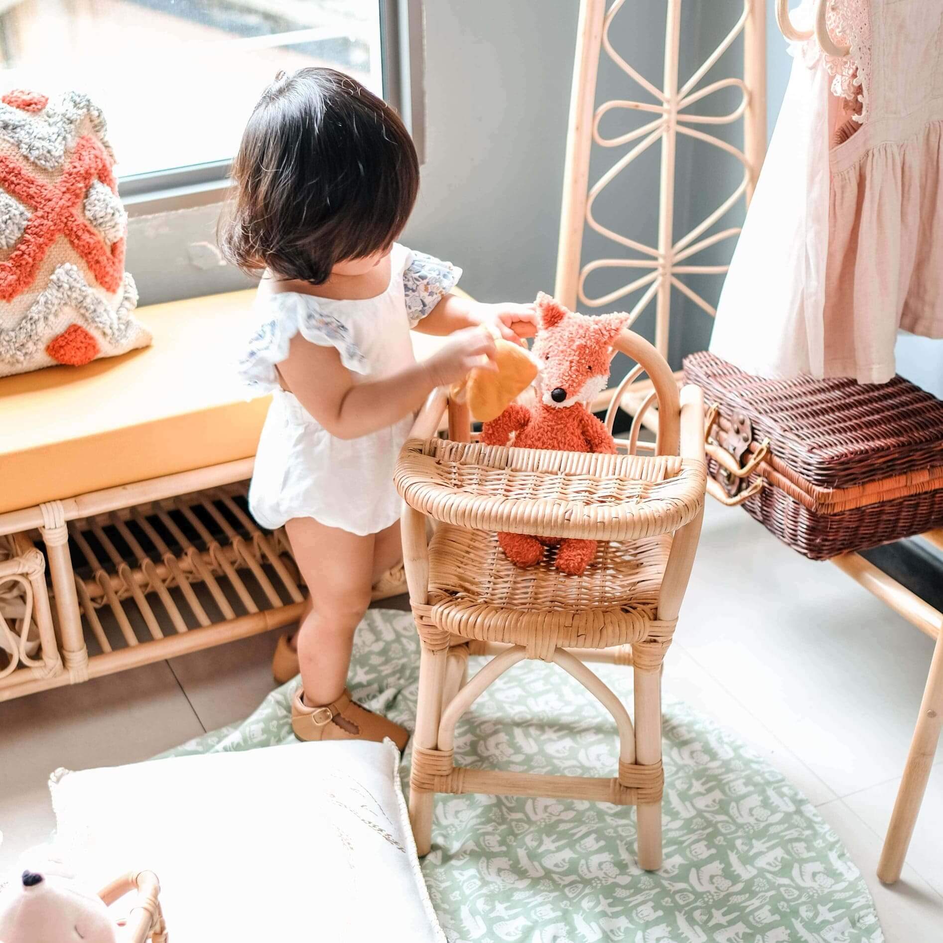 Child placing a doll onto the handcrafted rattan wicker Layla Doll High Chair by MOMIJI in a room with rattan furniture.