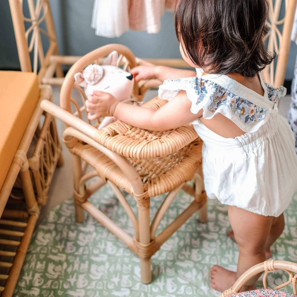 Front angled view of a child placing a doll onto the handcrafted rattan wicker Layla Doll High Chair by MOMIJI in a room with rattan furniture.