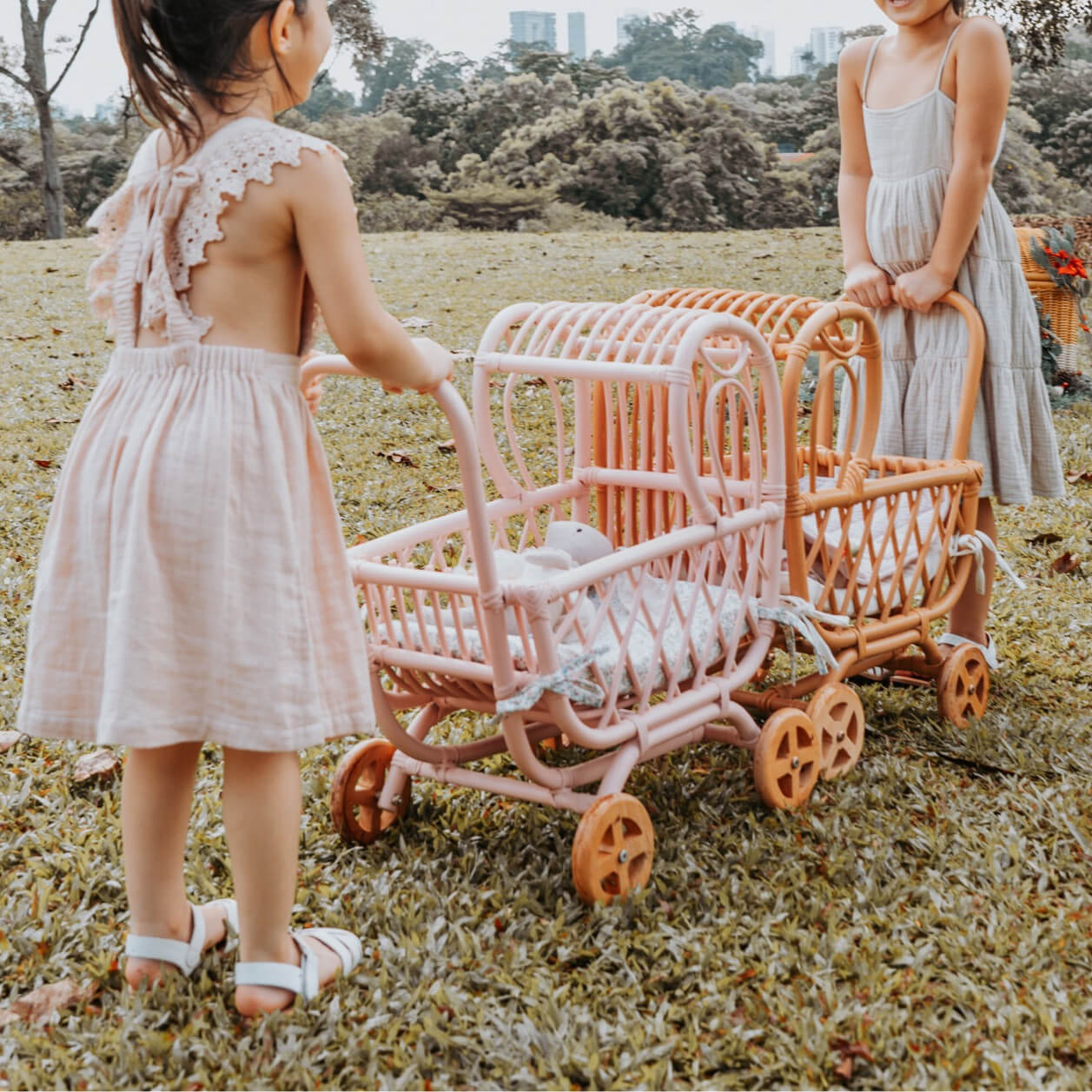 Two children playing with the handwoven rattan Layla Toy Stroller by MOMIJI in pink and natural versions with toy inside in a park setting.