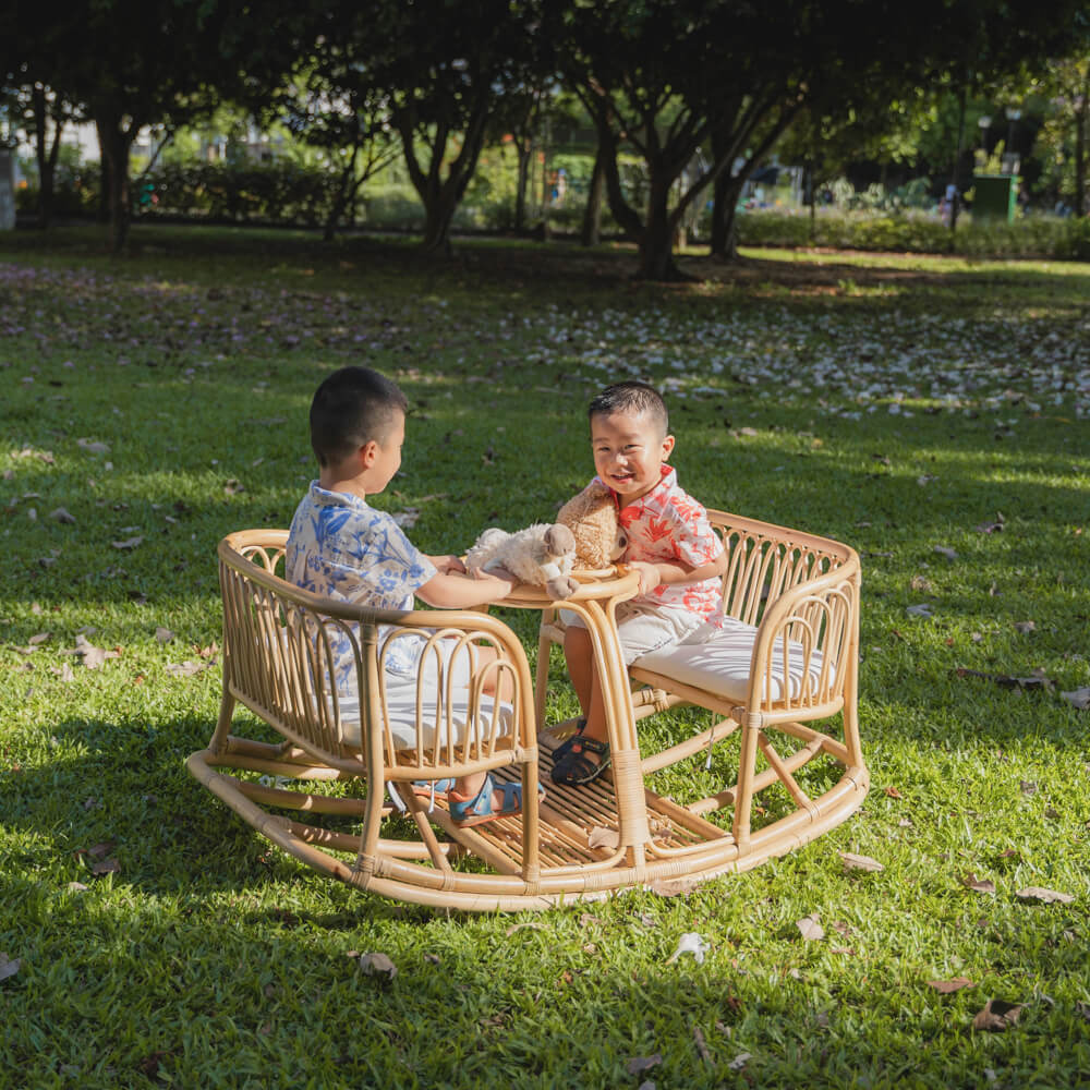 Two smiling children sitting and playing on the Leon Rocking Bench by MOMIJI with dolls in an outdoor garden.