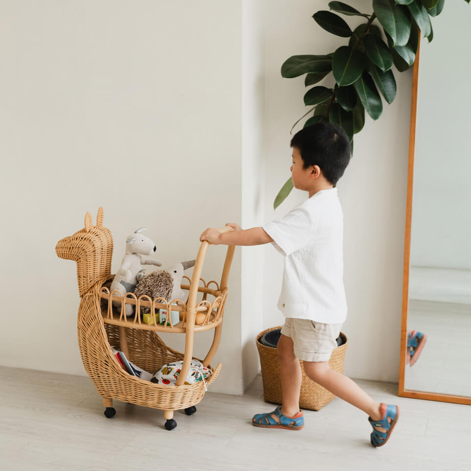 Child pushing a wicker Llama Craft Trolley filled with stuffed animals in a cozy room with indoor plants and a mirror.