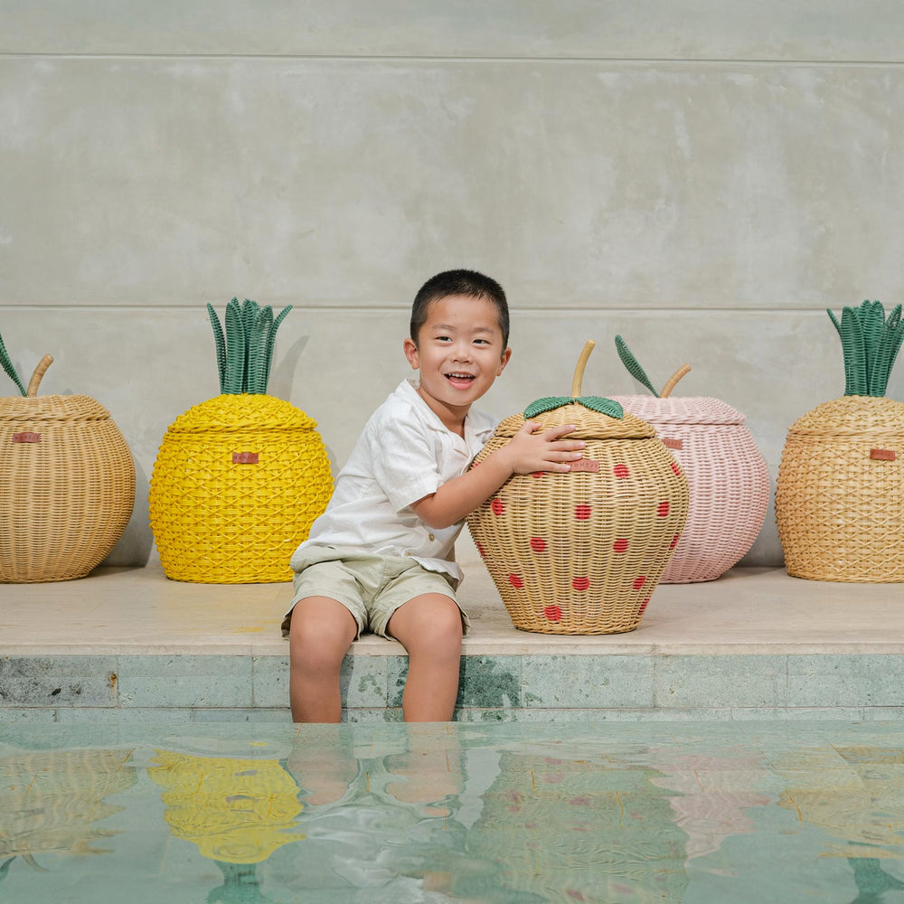 Child sitting by a pool holding a Strawberry Rattan Storage Basket with fruit designed rattan storage basket collection in the background.