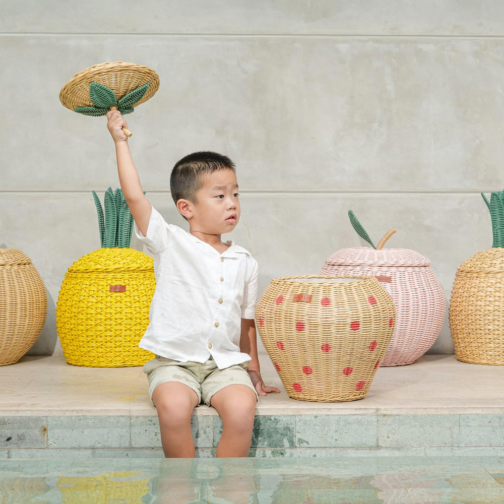 Child holding a woven rattan basket lid by a pool with fruit designed rattan storage basket collection in the background.