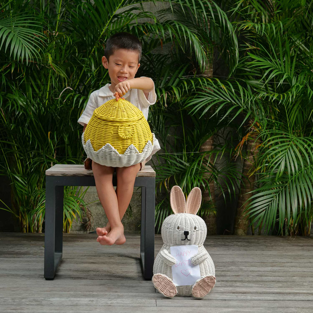 Child holding a Chick Rattan Storage Basket with Bunny Rattan Storage Basket by MOMIJI on the ground with plants on the background.