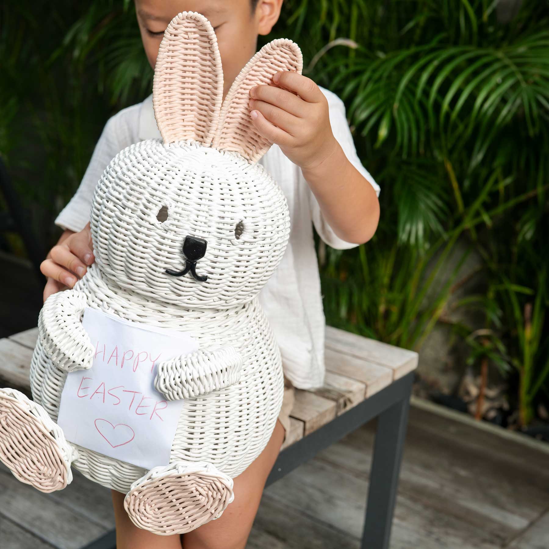 Child holding a Bunny Rattan Storage Basket by MOMIJI with a 'Happy Easter' sign outdoors.