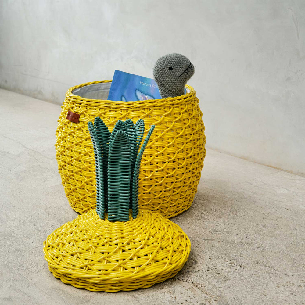 Side view of an opened yellow Pineapple Rattan Storage Basket with a book and a toy inside the basket, lid on the floor.