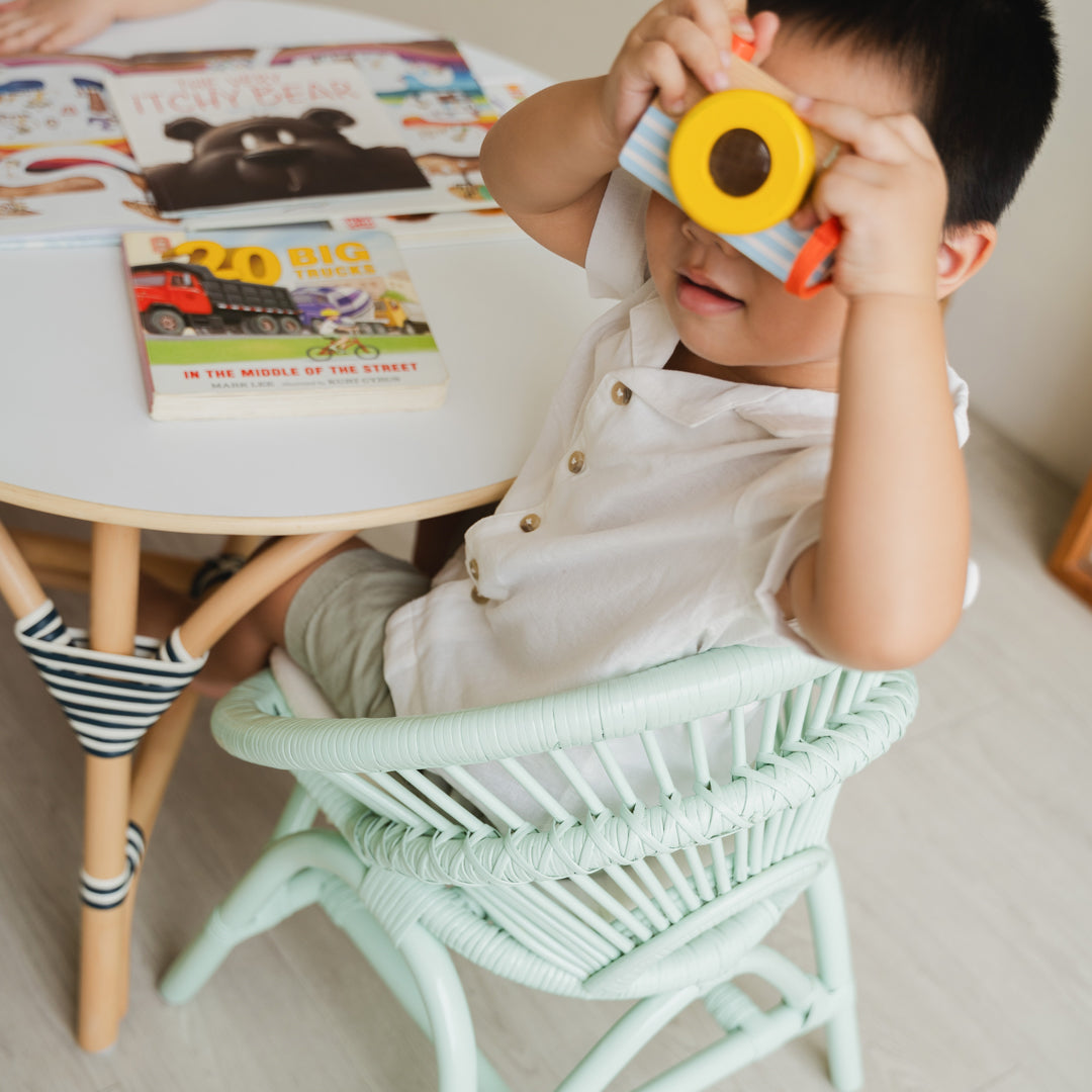 Child playing with a toy camera while sitting on the handcrafted Mint Green rattan Maya Kids Chair by MOMIJI  with books at an Aulia Kids Table, supporting independent and comfortable play in a playful children's room.