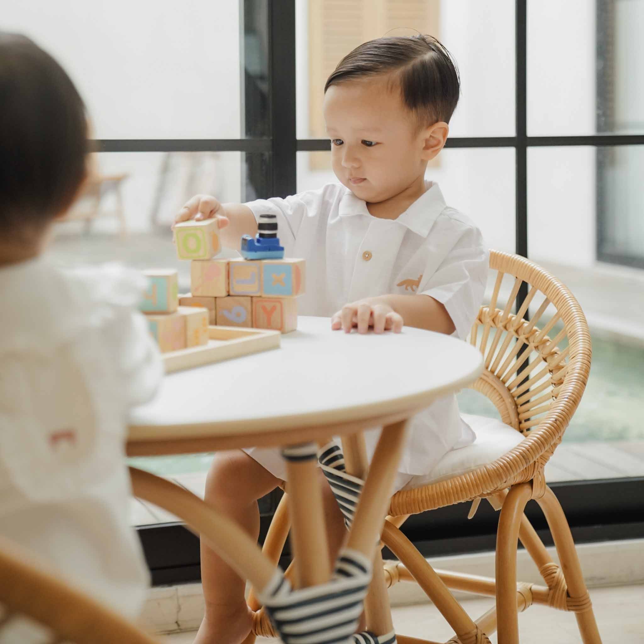 A child sits on a handcrafted natural rattan Maya Kids Chair by MOMIJI at a matching Aulia Kids Table, stacking colourful wooden blocks in a playful indoor setting.