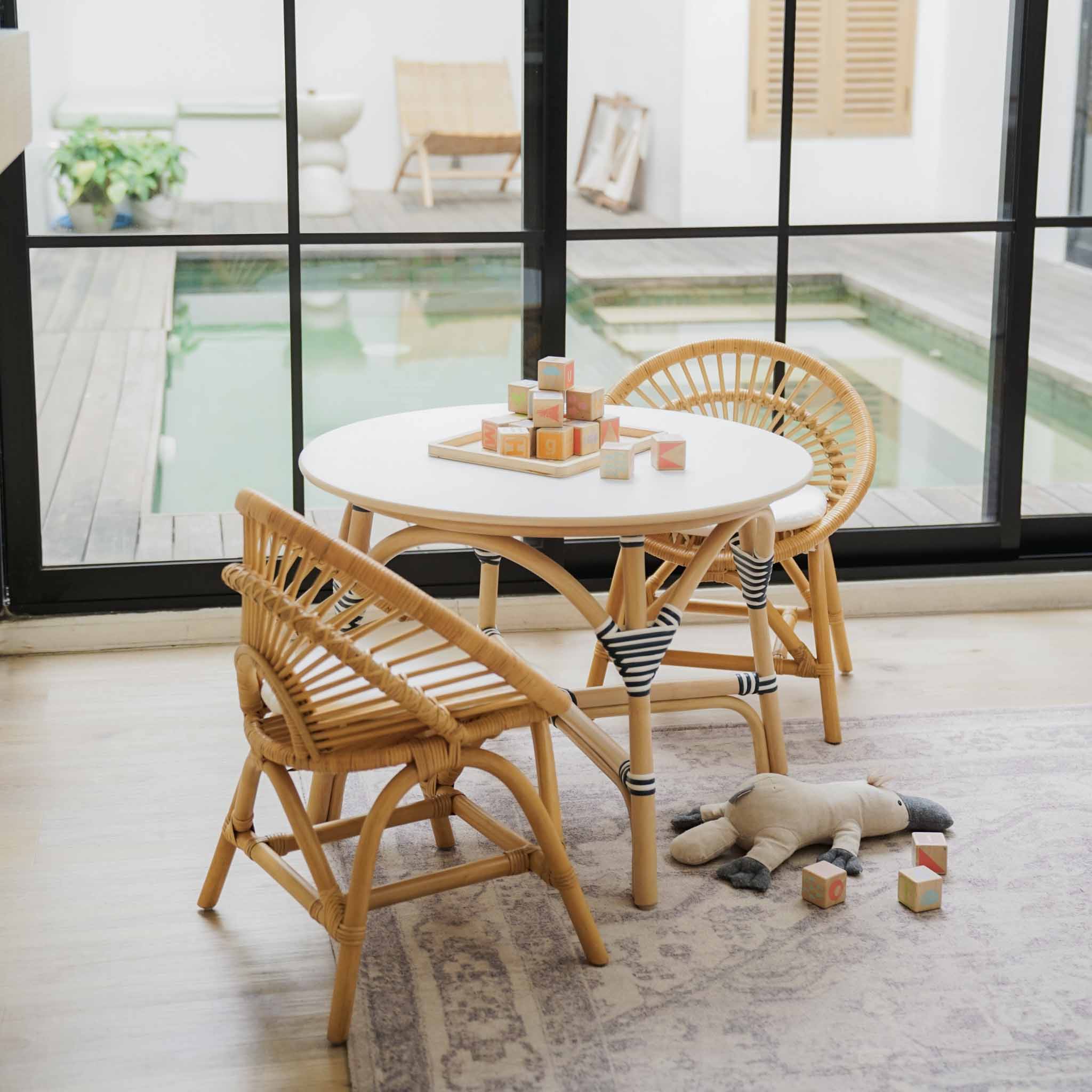 A children's dining set featuring an Aulia Kids Table and two handcrafted natural rattan Maya Kids Chairs by MOMIJI with black-and-white striped cushions, styled in a bright room with a large window overlooking a pool.