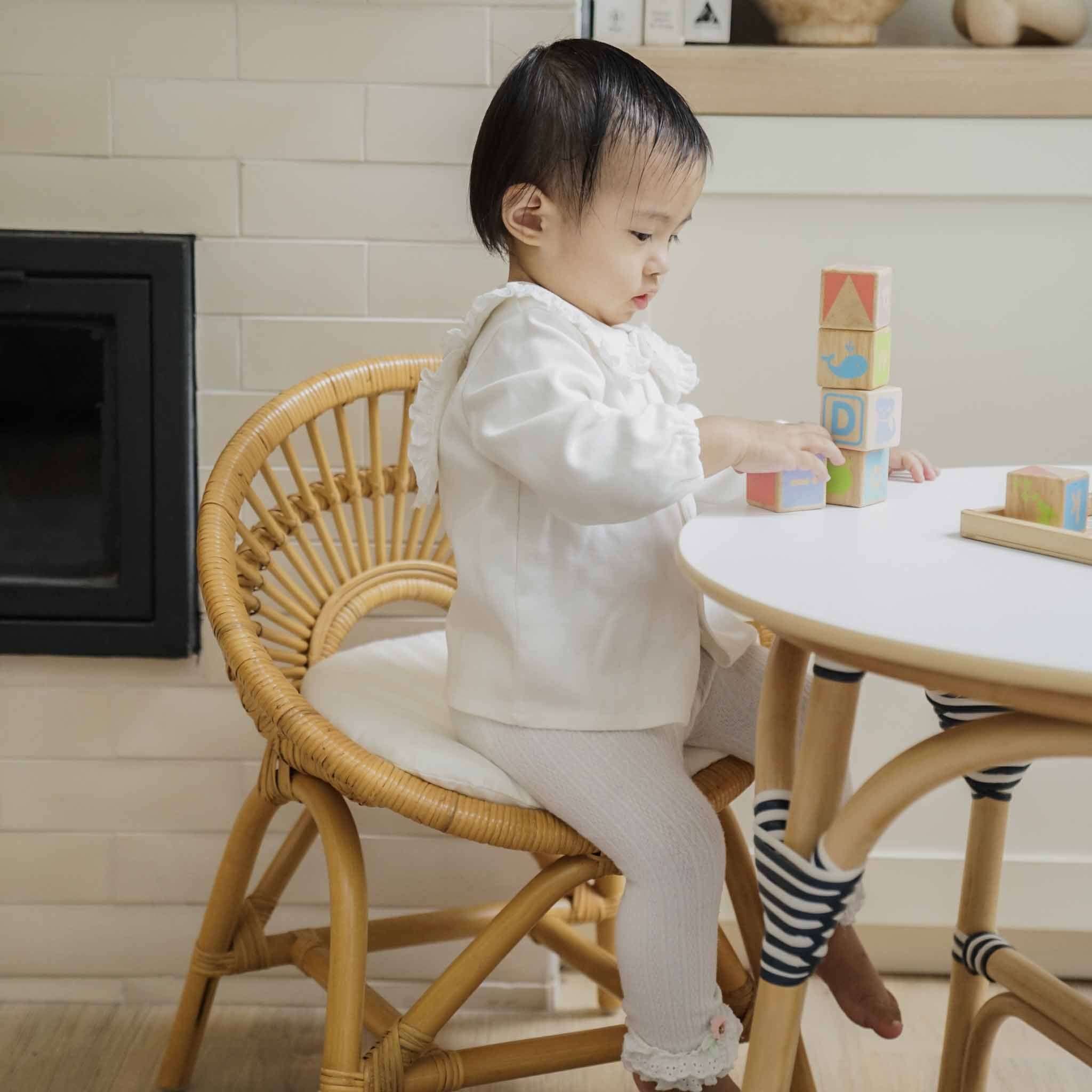 A toddler sits on a handcrafted natural rattan Maya Kids Chair by MOMIJI at a matching Aulia Kids Table stacking colourful wooden blocks in a room with a light tiled wall.
