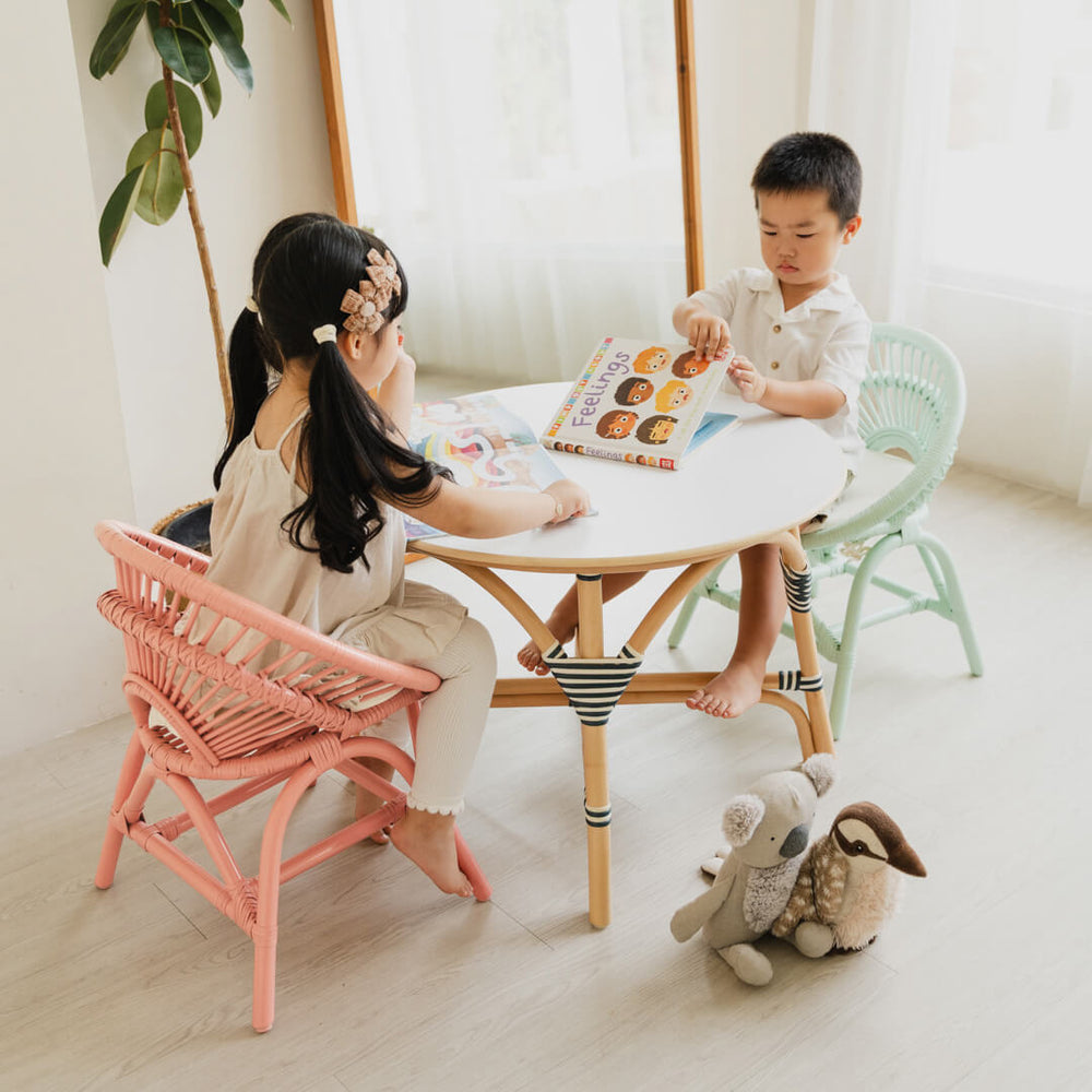 Two children sitting on Maya Kids Chairs peach pink and mint green by MOMIJI reading books in a room with a mirror and plants, with toys on the floor.