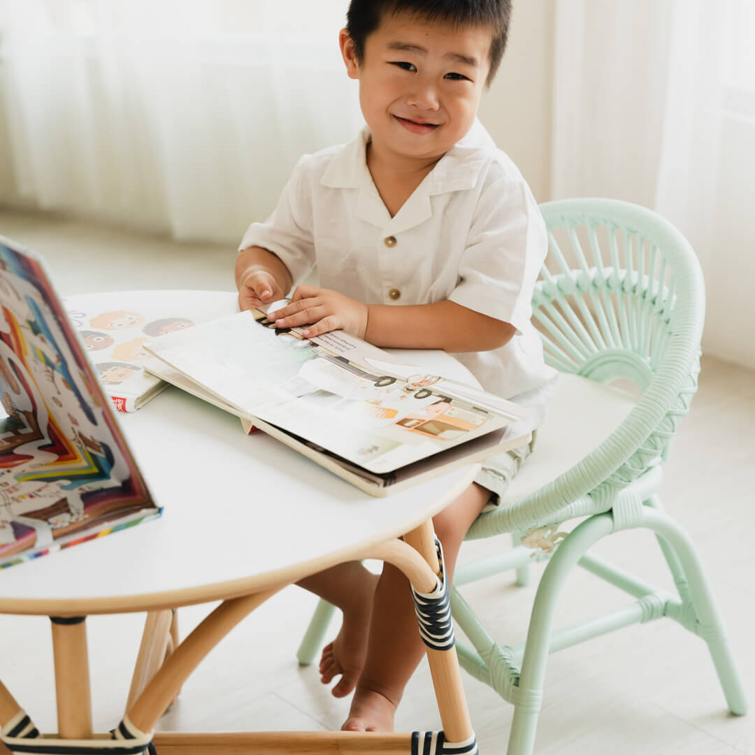 Smiling child sitting on the Maya Kids Chair by MOMIJI reading a book at a table.