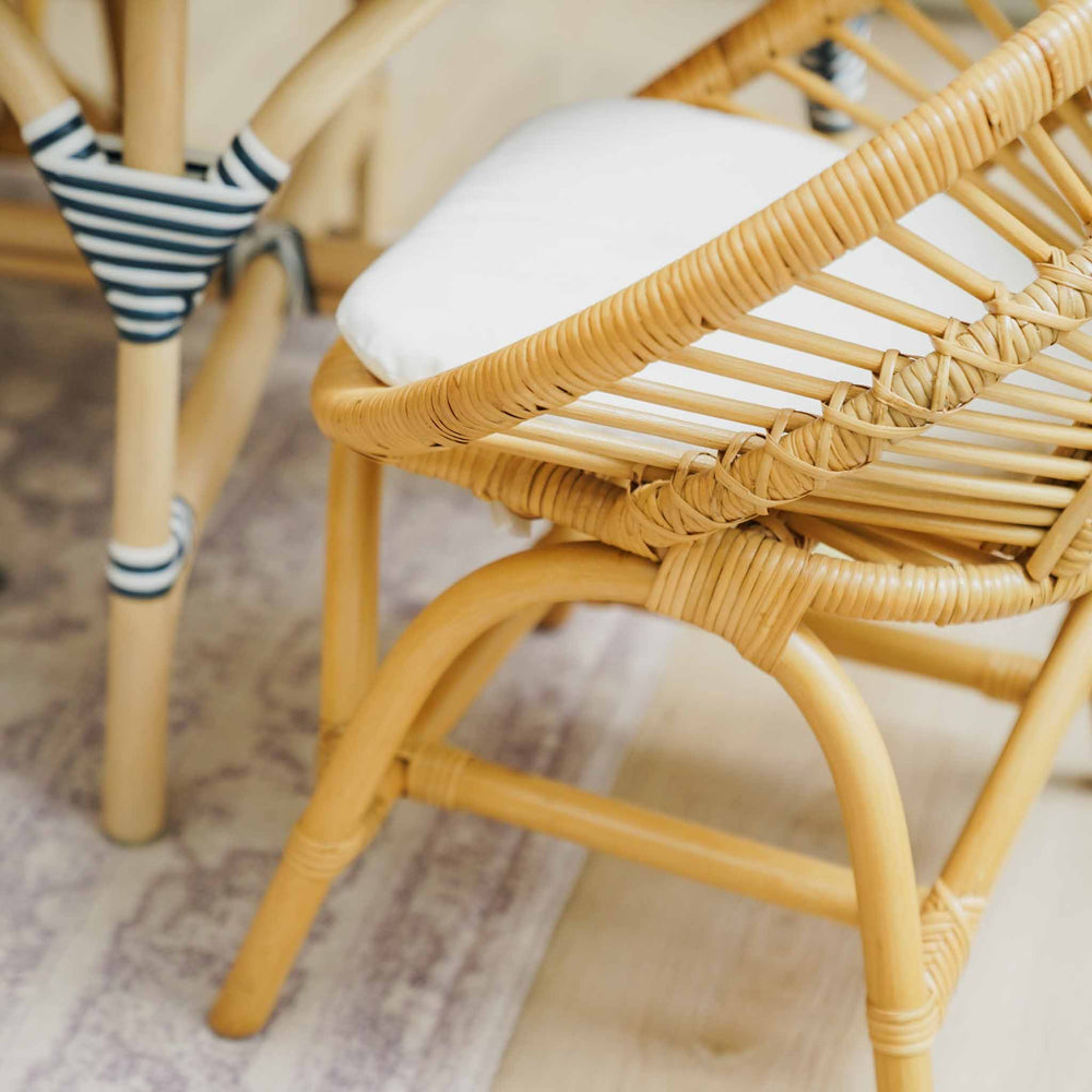 Close-up of the handcrafted natural rattan Maya Kids Chair by MOMIJI, highlighting its comfortable removable white cushion and smooth woven rattan details in a softly lit indoor setting with a patterned rug and another chair in the background.