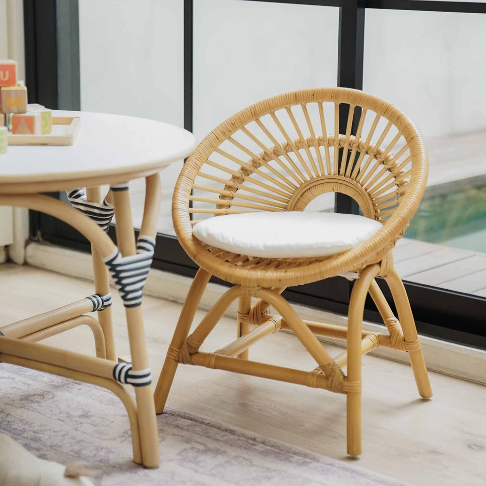Front view of the handcrafted rattan Maya Kids Chair by MOMIJI in natural color with a removeable white cushion, styled in a room with a rattan table.