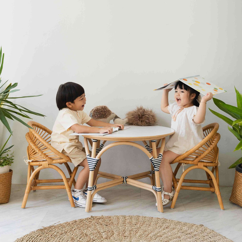 Two children sitting on the Maya Kids Chair by MOMIJI laughing and holding books at a table in a room with plants.