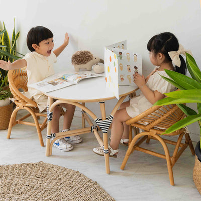 Child sitting on the handcrafted rattan Maya Kids Chair by MOMIJI while playing and reading books at a table in a room with plants.