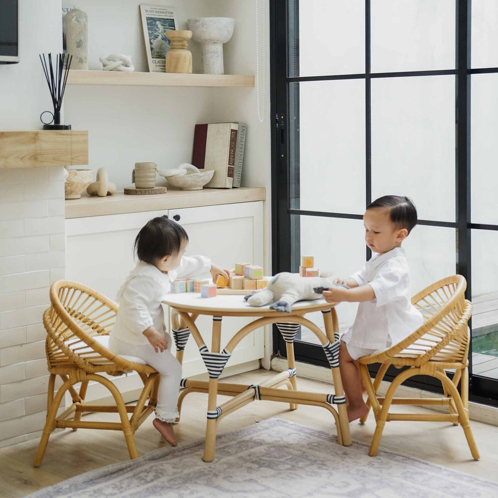 Two young children sitting on Natural handcrafted natural rattan Maya Kids Chairs by MOMIJI at a matching round rattan table in a bright, modern room with large black-framed windows.