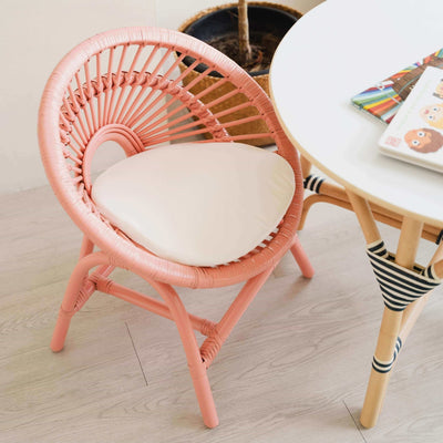 Tilted view of the handcrafted rattan Maya Kids Chair by MOMIJI in peach pink color with a white cushion, styled in a room with a rattan table.