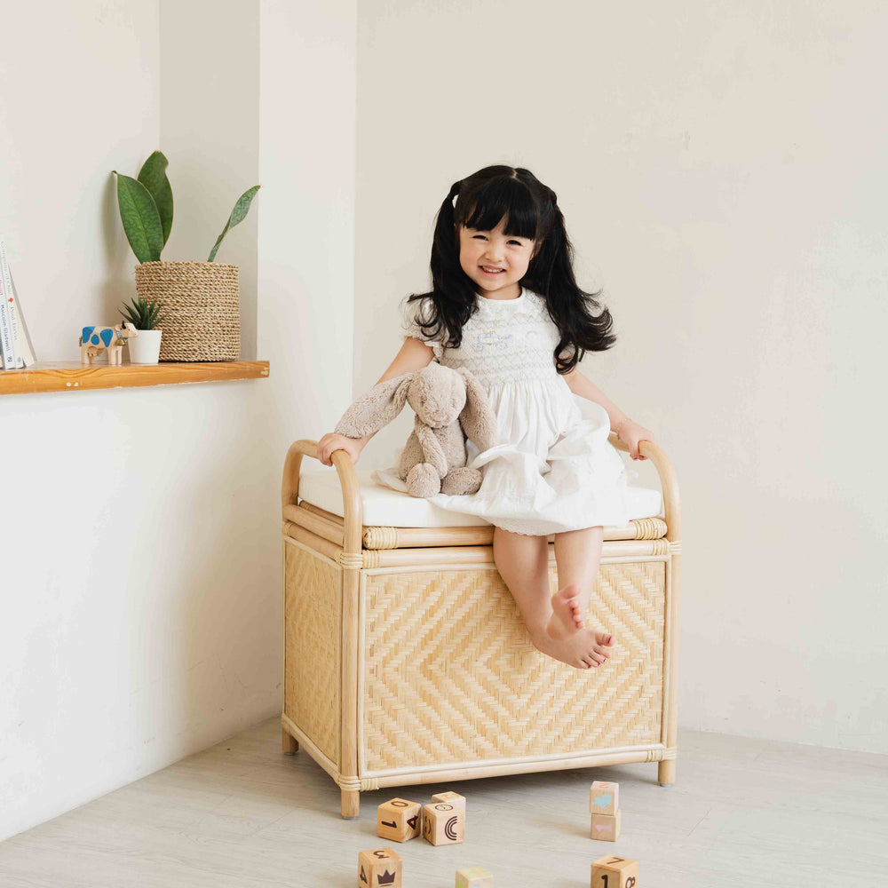 Smiling child sitting on the Mia Storage Bench by MOMIJI with a doll beside, styled in a cozy room with plants.