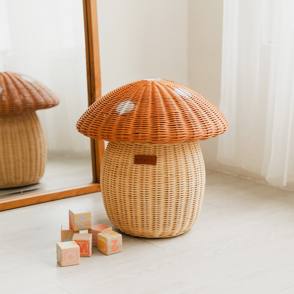 Mushroom Storage Rattan Basket by MOMIJI placed on a wooden floor next to a pile of toy blocks and a mirror behind, with a reflection of a similar basket in the mirror.