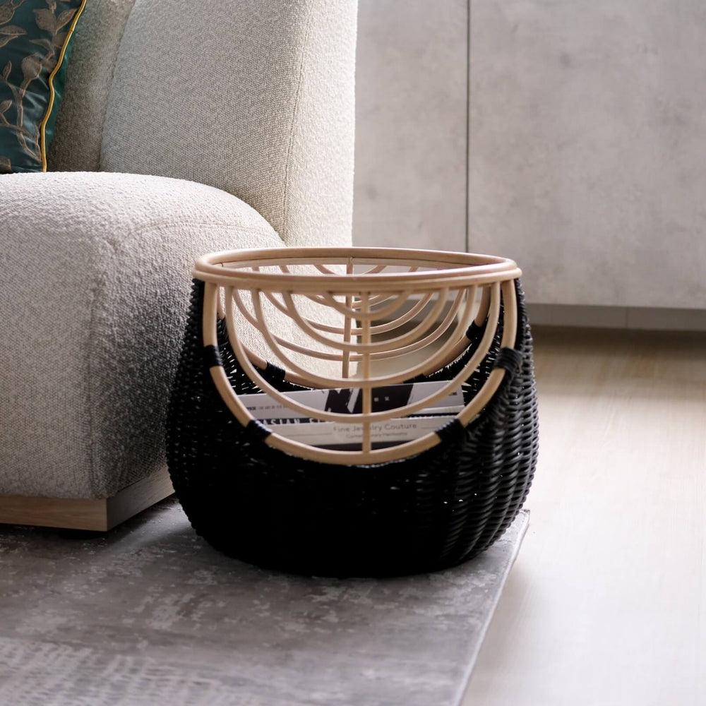 Side view of the handcrafted rattan wicker Nikko Basket by MOMIJI featuring black weaving with natural rattan accents, filled with books placed on a rug beside a sofa.