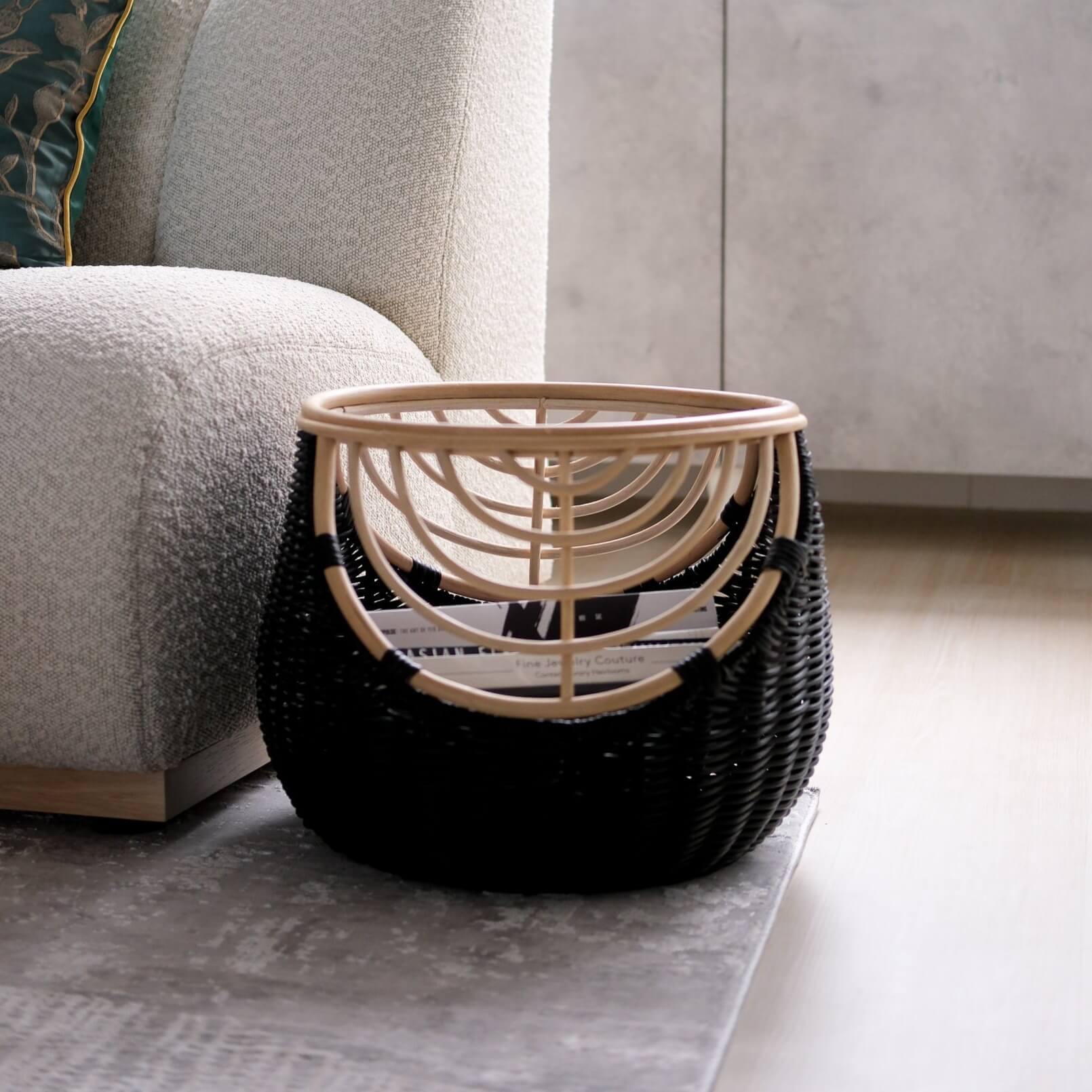 Side view of the handcrafted rattan wicker Nikko Basket by MOMIJI featuring black weaving with natural rattan accents, filled with books placed on a rug beside a sofa.