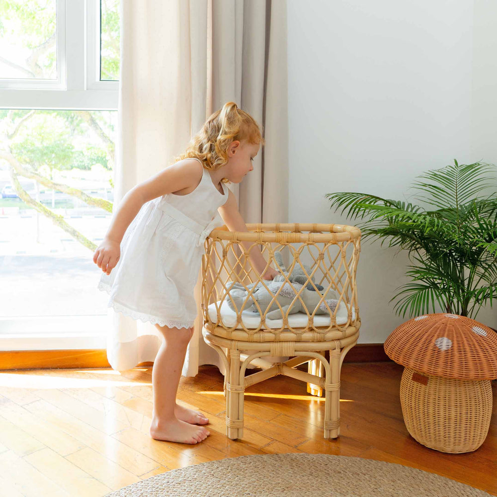 Child holding a doll inside the Olivia Doll Bassinet by MOMIJI in soft natural light, styled with plants and a rattan mushroom basket, featuring a fully handwoven rattan bassinet with white cushion.