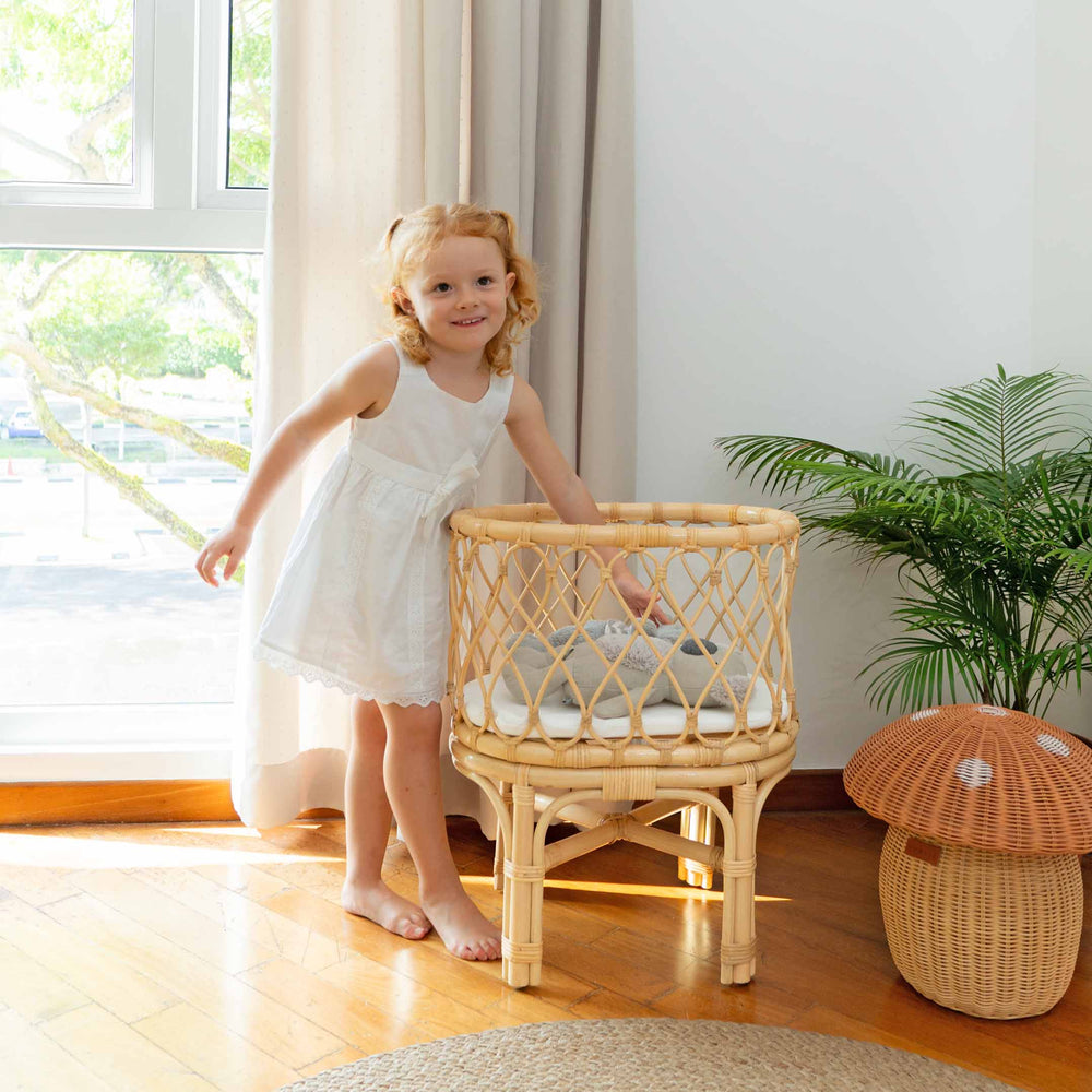 Smiling child playing beside the Olivia Doll Bassinet by MOMIJI in a room with plants and a rattan mushroom basket, featuring a fully handwoven rattan bassinet with white cushion.