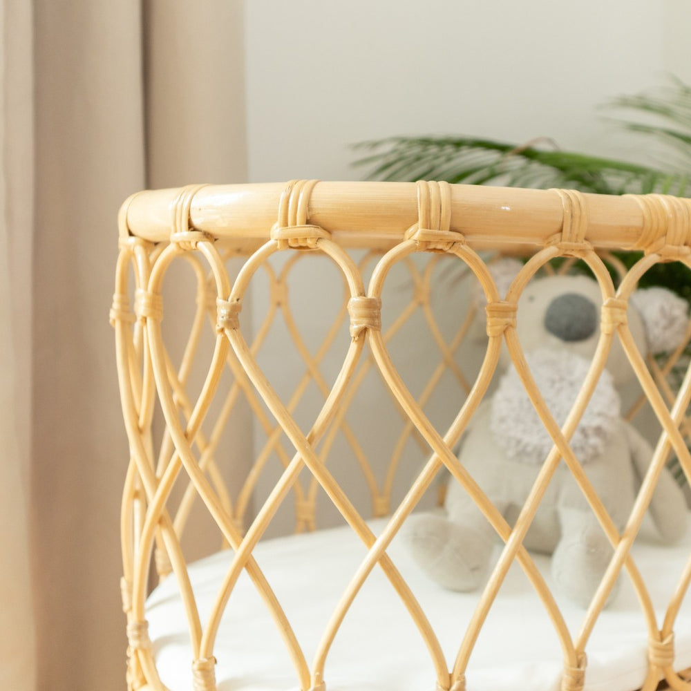 Side view of the Olivia Doll Bassinet by MOMIJI highlighting carved handwoven rattan detailing on the sides, with white cushion.