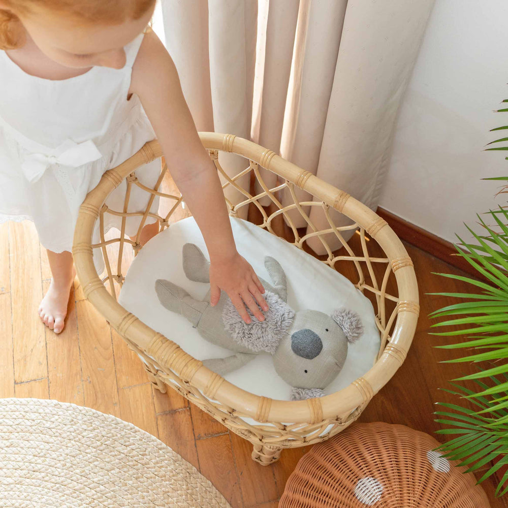 Top view of the Olivia Doll Bassinet by MOMIJI with a child gently touching a koala doll inside, featuring a fully handwoven rattan bassinet with white cushion.
