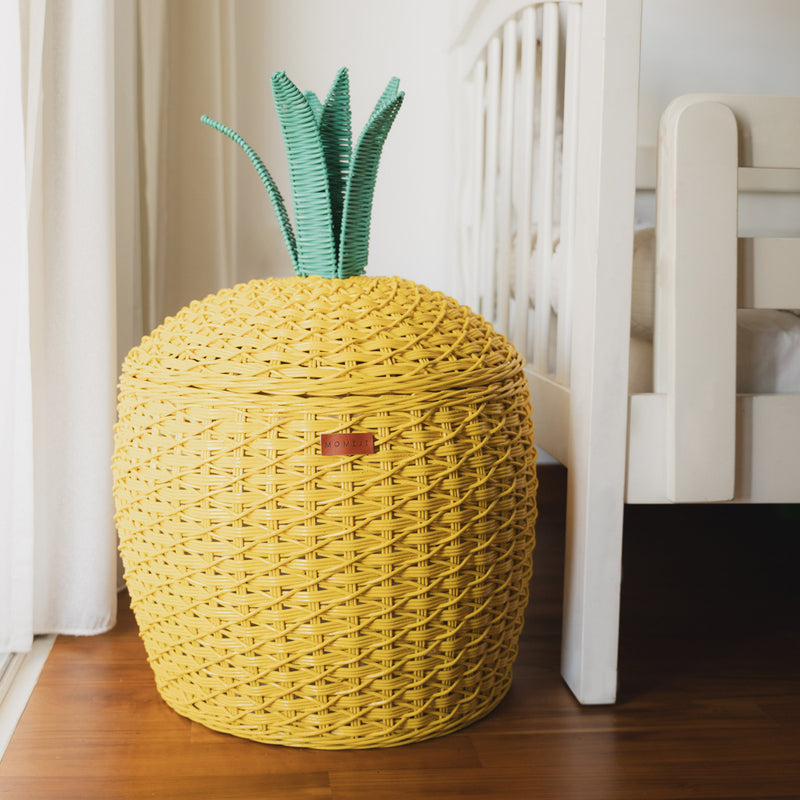 A yellow Pineapple Rattan Storage Basket placed on a floor next to a white bed frame.